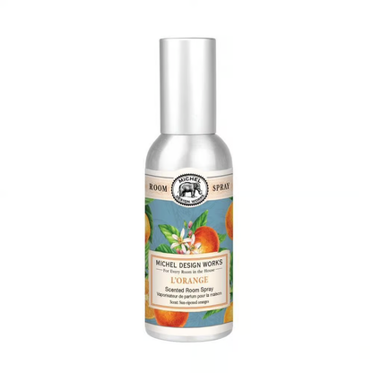A silver metal container of Michel Design Works L'Orange Scented Room Spray with a label featuring an orange and white design, and a printed sleeve in the background.