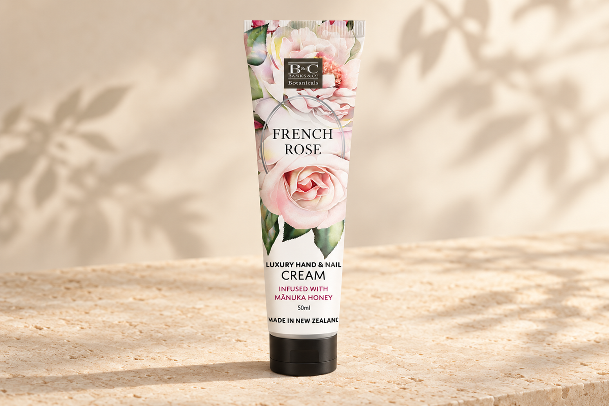 A squeeze tube of B&C French Rose Luxury Hand & Nail Cream stands upright on a textured beige stone surface with soft leaf shadows cast across a warm cream background.
