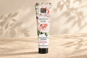 A squeeze tube of B&C French Rose Luxury Hand & Nail Cream stands upright on a textured beige stone surface with soft leaf shadows cast across a warm cream background.