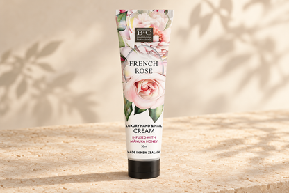 A squeeze tube of B&C French Rose Luxury Hand & Nail Cream stands upright on a textured beige stone surface with soft leaf shadows cast across a warm cream background.